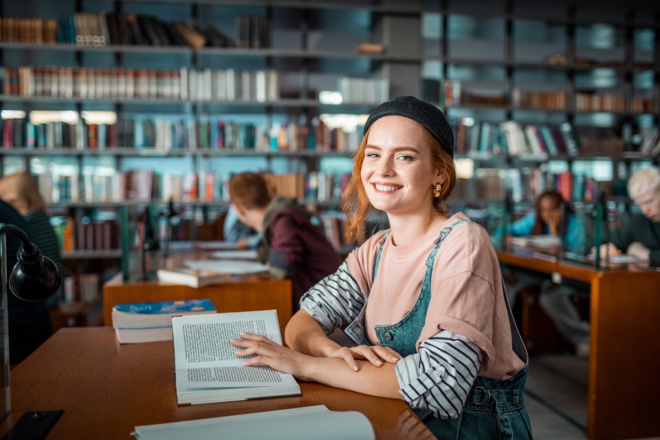 Young female student sitting at library table with open book smiling