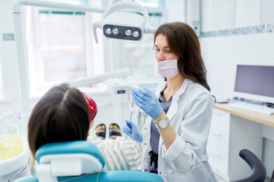 Female dentist wearing white coat and mask with patient in a chair in dental practice room