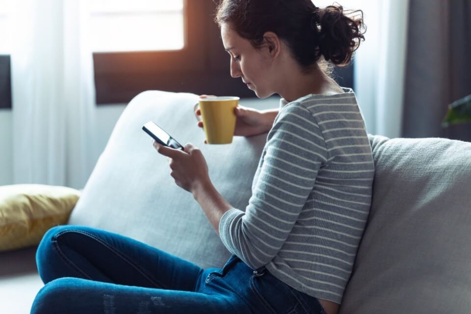 Young female sitting on sofa holding mug and phone