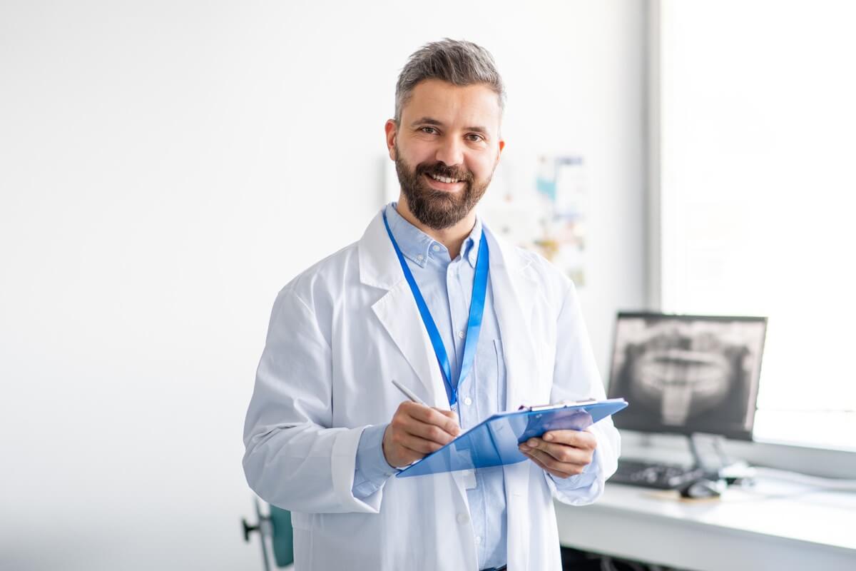 Male dentist in dental room wearing a white coat and holding a clipboard