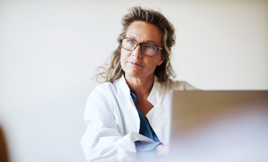 Female doctor sitting at desk wearing a white coat using a laptop