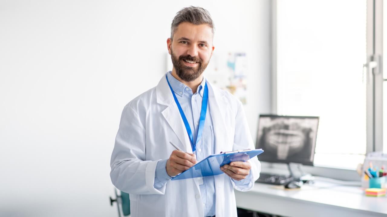 Male dentist in dental room wearing a white coat and holding a clipboard