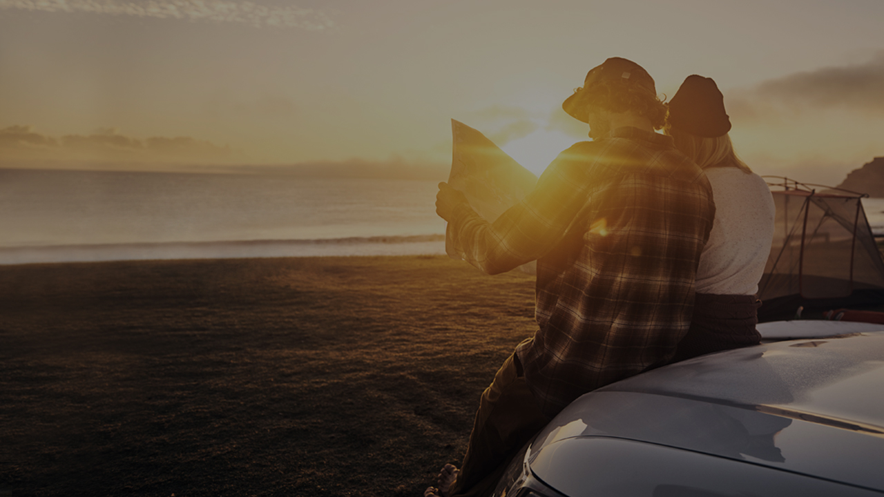 Couple sit on car looking at map while on the beach at sunset