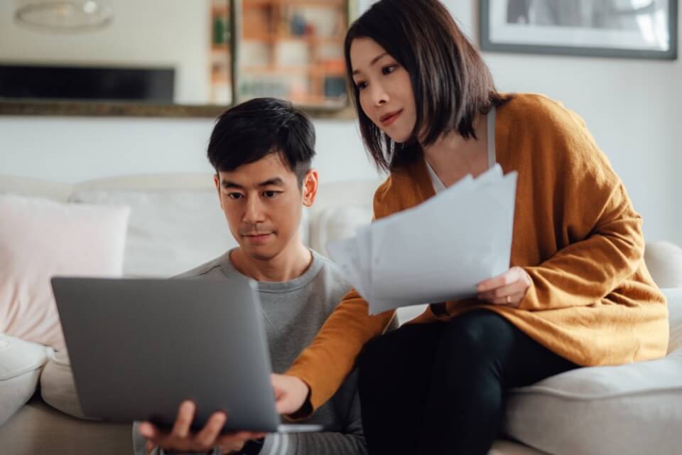 Young couple on sofa using laptop holding paperwork